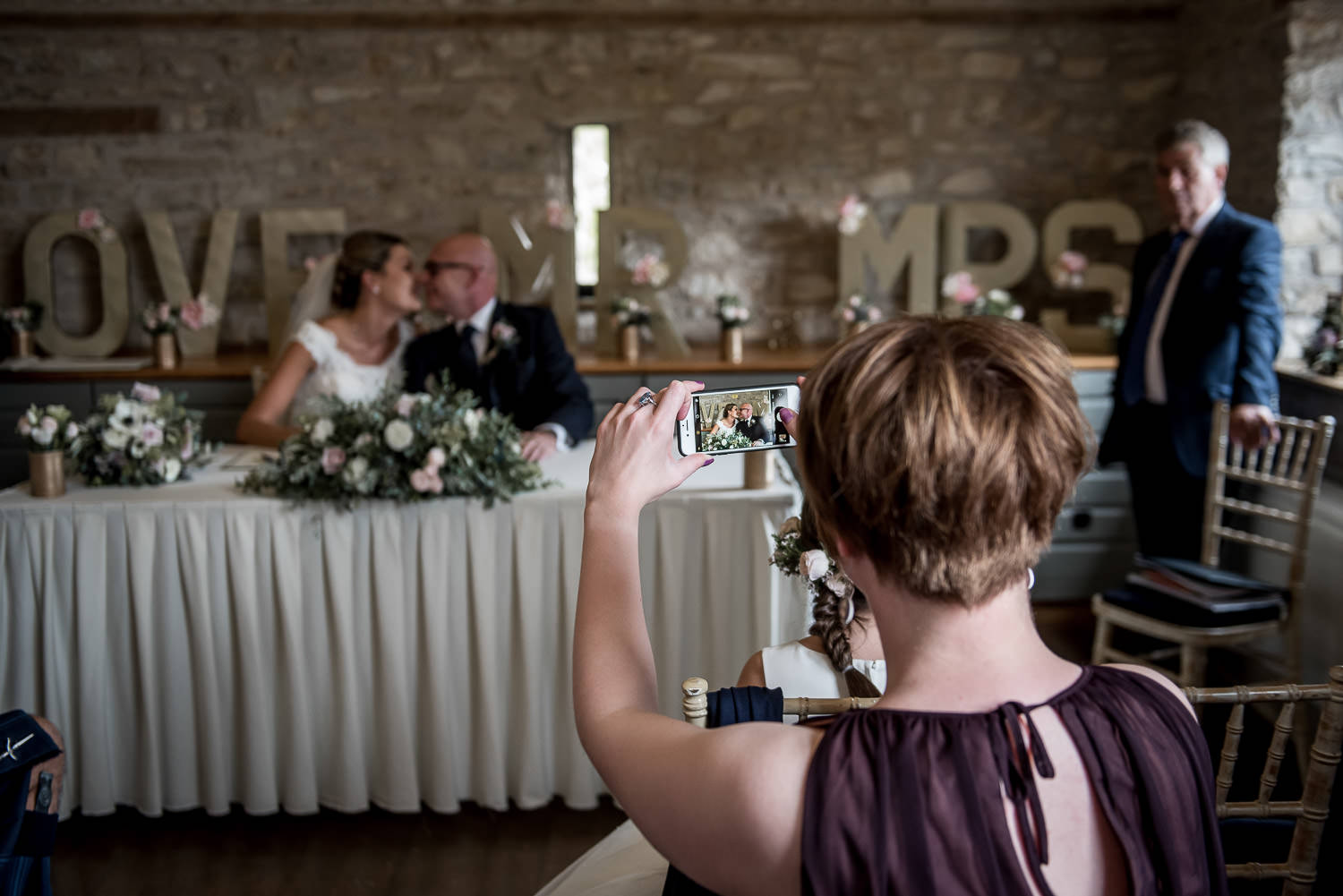 bridesmaid taking a photograph of the bride and groom kissing